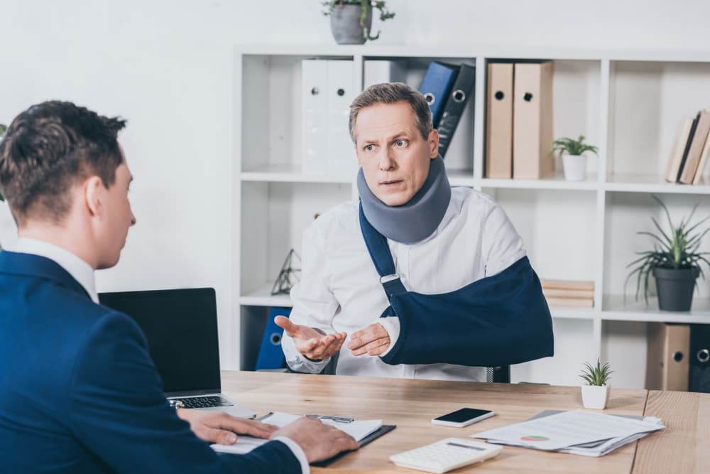 A middle-aged worker wearing a neck brace and with a broken arm sits at a table, discussing compensation with a businessman in a blue jacket in an office setting.