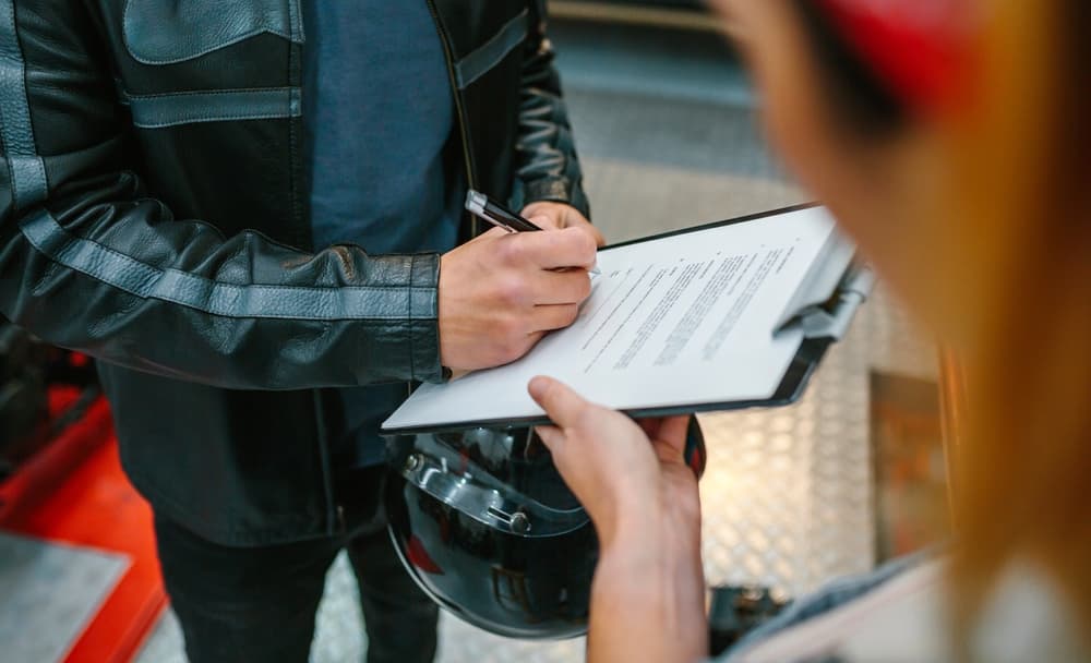 A biker, unidentifiable beneath a leather jacket and holding a helmet, is seen signing an insurance policy. This signifies his intent to collect his repaired motorcycle from the workshop.